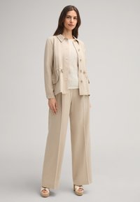 Blazer beige con vita regolabile e tasche, abbinato a una leggera maglietta di crema texturizzata e pantaloni beige a gamba larga. Sandali con zeppa.