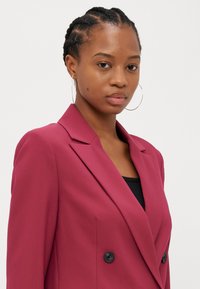Blazer doppiopetto di un rosso intenso, con silhouette sagomata, revers a scaglione e bottoni neri. Tessuto liscio con una finitura strutturata.