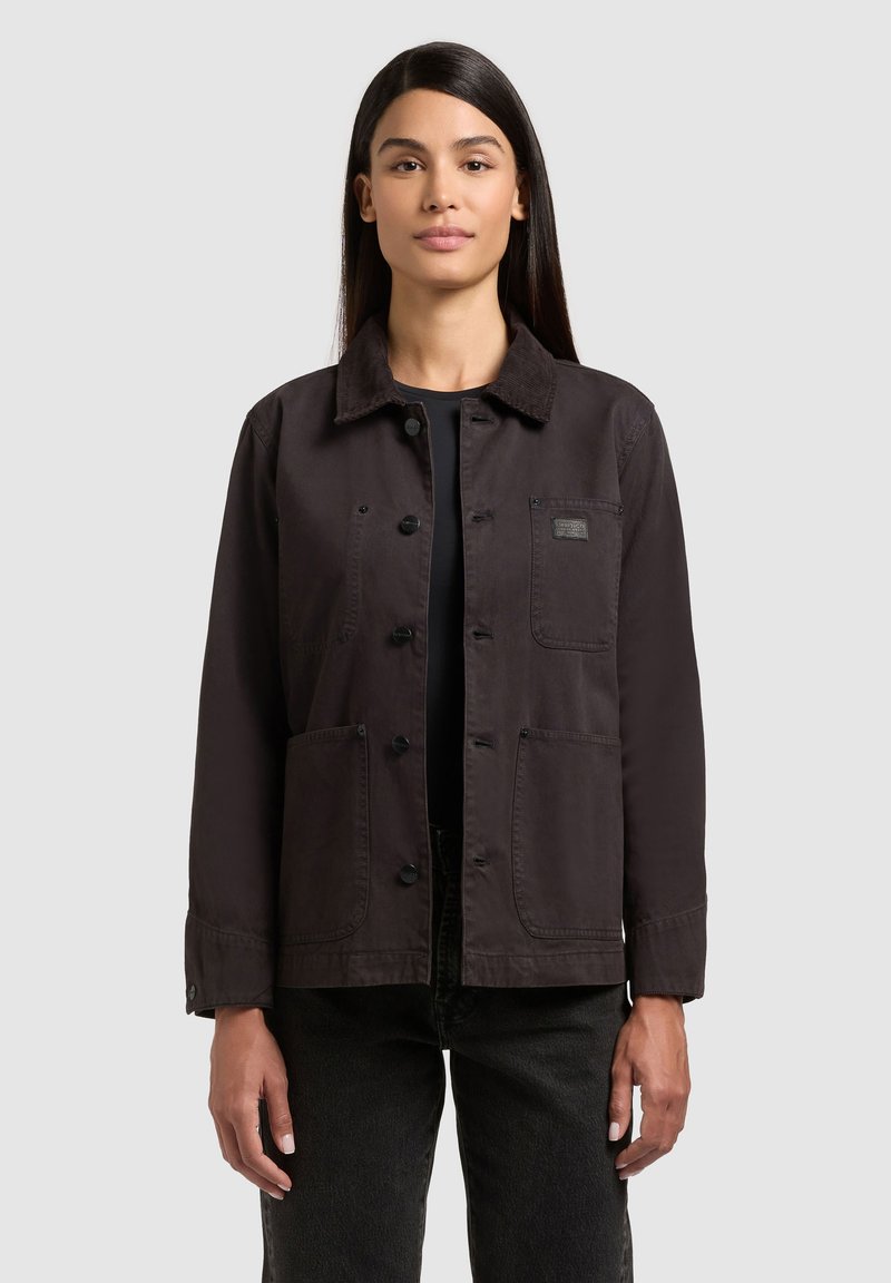 Jeune femme aux longs cheveux foncés portant une veste marron foncé boutonnée sur un haut noir et un pantalon sombre, debout devant un fond uni.