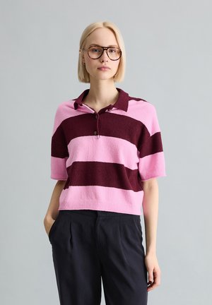 Blonde Frau mit übergroßen braunen Brillen, kurzärmeligem pink- und burgunderrot gestreiftem Poloshirt und schwarzer Hose, stehend vor grauem Hintergrund.