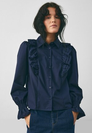 Námořnická modrá košile na knoflíky s nařasenými detaily na přední straně a ramenech, má nabírané rukávy se smockovanými manžetami a rovný lem.