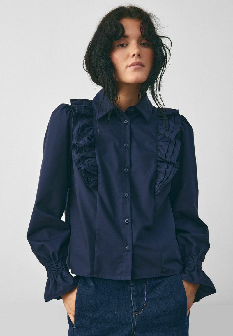 Tamsiai mėlyna mygtukinė marškinėliai su raukšlėmis priekyje ir ant pečių, turintys pūstus rankoves su susiūtomis rizelėmis ir tiesiu apdailu.