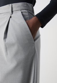 Pantalons gris texturés avec des plis et des poches à main. Le tissu semble léger avec un motif subtil. Vue rapprochée de la taille et de la main.