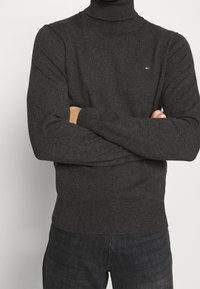 Maglione a collo alto grigio scuro con collo e polsini a costine, dotato di un piccolo logo sul petto, abbinato a jeans scuri.