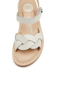 Sandale en cuir blanc avec un design de sangle tressée, une semelle plate et une sangle de cheville dotée d'une boucle en métal. Semelle intérieure beige douce avec des coutures visibles.