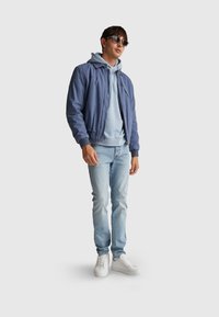 Giacca bomber azzurro chiaro, felpa grigia con cappuccio, jeans azzurro chiaro e sneakers bianche. Il modello indossa occhiali da sole scuri. Design minimalista, stile casual.