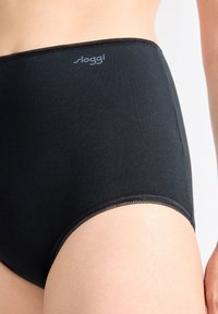 Ingrandimento di una persona che indossa mutandine in cotone nere a vita alta con il logo "sloggi" sulla cintura, mostrando parte dell'anca e della mano.