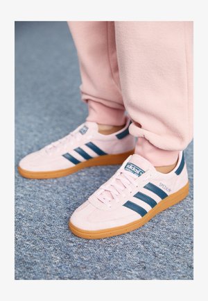 Sneakers de camurça rosa claro com riscas de pele azul escura e sola de borracha. Características incluem atacadores planos e marcação no lado e na língua.