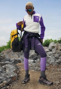 Pulover din fleece alb și violet, colanți violeți, rucsac galben și negru, ochelari de soare și bocanci pe un teren stâncos.