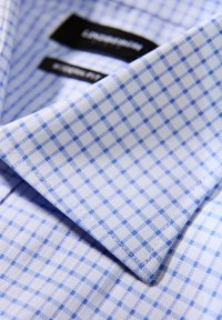 Camicia azzurro chiaro a quadri con colletto alla francese, caratterizzata da una trama intrecciata e motivi a griglia di colore blu scuro a contrasto. Etichette visibili all'interno del colletto.