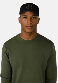 Grön tröja med rund halsringning, strukturerad tyg; svart basebollkeps med en broderad bokstav "C." Modellen visar ett neutralt uttryck.