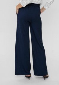 Pantalon large bleu marine avec taille élastique, doté de poches latérales et d'une texture lisse. Assorti à des talons noirs élégants.