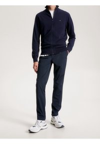 Sudadera con cremallera azul marino, pantalones oscuros y zapatillas blancas. Textura suave, diseño ajustado, logo en el pecho, estilo minimalista y tonos neutros.