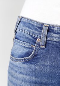 Pantalones vaqueros de denim con un lavado medio, diseño clásico de cinco bolsillos, corte recto, con costuras doradas y acentos de hardware en bronce.
