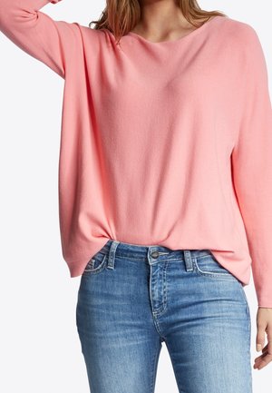 Kobieta w luźnym różowym swetrze z długim rękawem, włożonym do wysokich dżinsów z zapięciem na guzik i suwak.