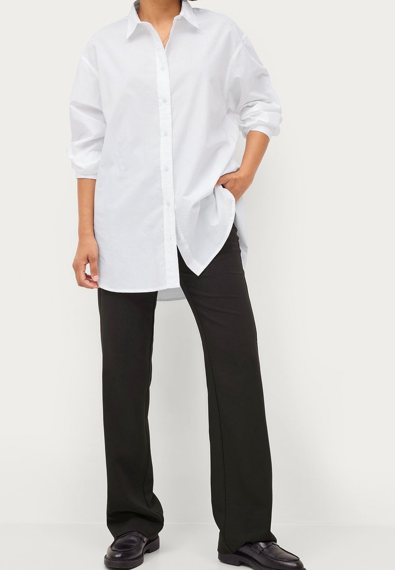 Personne portant une longue chemise blanche boutonnée avec manches retroussées, pantalon droit noir et chaussures noires, debout avec une main dans la poche.