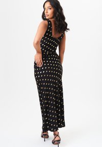 Black maxi dress with gold oval polka dots, sleeveless design, fitted silhouette, and a scoop back. Worn with strappy black heels.