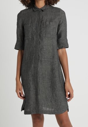 Kvinde iført en knælång, mørkegrå skjortekjole med korte ærmer, krave, knapstolpe foran og to brystlommer.