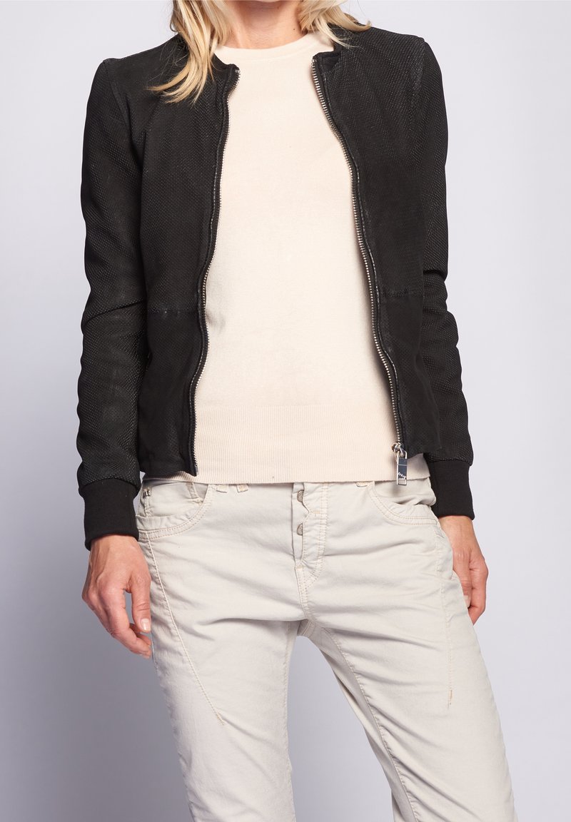 Svart texturerad zip-up bomberjacka med rund hals och ribbade ärmar, bärs över en ljusbeige tröja och ljusa byxor.