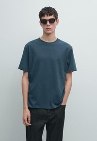 T-shirt di cotone verde petrolio scuro con collo rotondo e maniche corte, abbinata a jeans scuri in denim e occhiali da sole rettangolari neri.