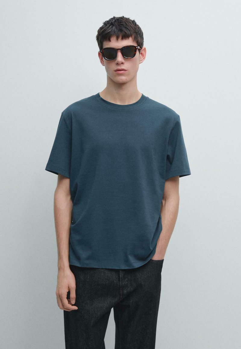 T-shirt di cotone verde petrolio scuro con collo rotondo e maniche corte, abbinata a jeans scuri in denim e occhiali da sole rettangolari neri.