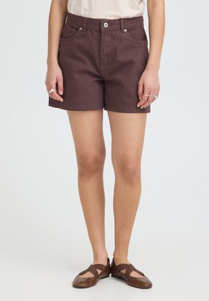 Žena oblečená v tmavohnedých rifľových šortkách, béžovom topu a hnedých plochých topánkach s elastickými pásikmi, stojaca pred jednoliatym svetlým pozadím.