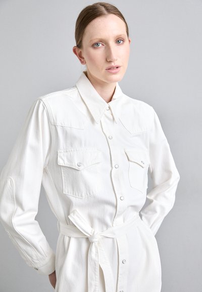 Balta marškiniai su atviru apykaklės kirpimu, dviem krūtinės kišenėmis ir prilaisvinta juosmens detale. Pagaminti iš tekstūruoto audinio, laisvo silueto.