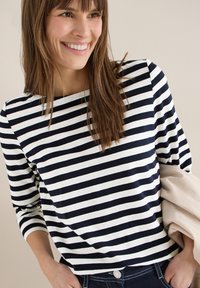 Femme souriante aux longs cheveux bruns portant un haut rayé noir et blanc et tenant une veste beige, debout devant un fond clair uni.