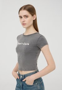 Maglietta corta grigia realizzata in tessuto morbido, con maniche corte. Il testo frontale recita "Non mi interessa" in bianco, con un design aderente.