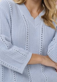 Hellblauer, gestrickter Pullover mit V-Ausschnitt, grober Textur und offenem Muster entlang der Ärmel und des Körpers.
