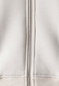 Helle beige Zip-Hoodie mit einem metallischen Reißverschluss in der Mitte, aus weichem Baumwollstoff, mit gerippter Saum und glatter Textur.