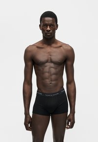 Μυώδης άνδρας στέκεται όρθιος φορώντας μαύρα μποξεράκια Tommy Hilfiger μπροστά σε απλό λευκό φόντο.