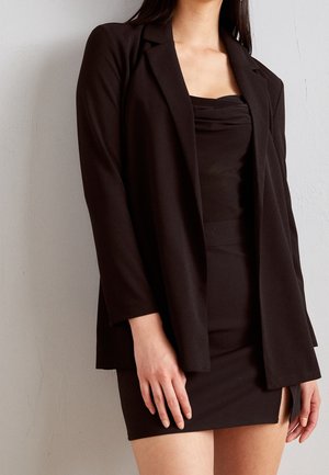 Femme portant un blazer noir par-dessus un top noir sans bretelles et une mini-jupe ajustée, debout devant un mur clair uni.
