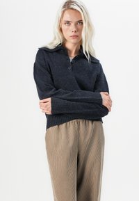 Ciemnoszary sweter robiony na drutach z kołnierzykiem, dwoma guzikami i ściągaczami; zestawiony z luźnymi, paskowanymi beżowymi spodniami.
