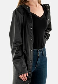 Cappotto lungo impermeabile nero con cappuccio, bottoni a pressione e una texture liscia. Indossato sopra un top nero e jeans in denim blu.