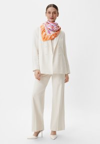 Biała, teksturowana marynarka na jasnej koszulce na ramiączkach, zestawiona z kremowymi spodniami o szerokich nogawkach i kolorowym jedwabnym szalem w pomarańczowe i różowe wzory.
