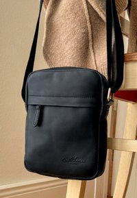 Černá kožená crossbody taška s plochým designem, zipovým uzávěrem, přední kapsou a nenápadným embosovaným logem. Odepínatelný popruh pro snadné nošení.