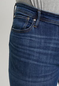 Κοντινό πλάνο ενός ατόμου που φορά σκούρα μπλε τζιν με πορτοκαλί ραφές και μια γκρι μπλούζα τυλιγμένη στη μέση.