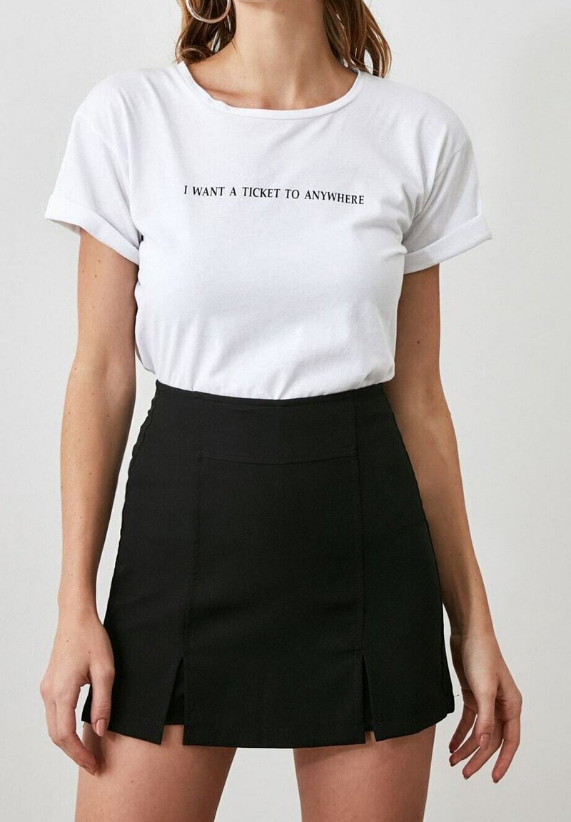 Mujer con una camiseta blanca que lleva el texto "QUIERO UN BOLETO A CUALQUIER LUGAR" y una falda mini negra con aberturas frontales.