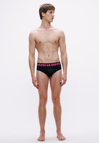 Mustad ujumispüksid erksate roosade "DSQUARED2" logodega vöökohal, slim-fit lõikega, pehmest materjalist ja siledapinnalise tekstuuriga, mida kannab seisv modell.