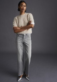 Donna con capelli corti e scuri che indossa un maglione beige peloso, jeans larghi grigio chiaro e scarpe argentate a punta, in piedi con le braccia incrociate.