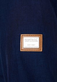 Patch en cuir marron avec le logo et le texte Regatta Great Outdoors "EST. 1981 FOR EVERY ADVENTURE" cousu sur un tissu bleu foncé.