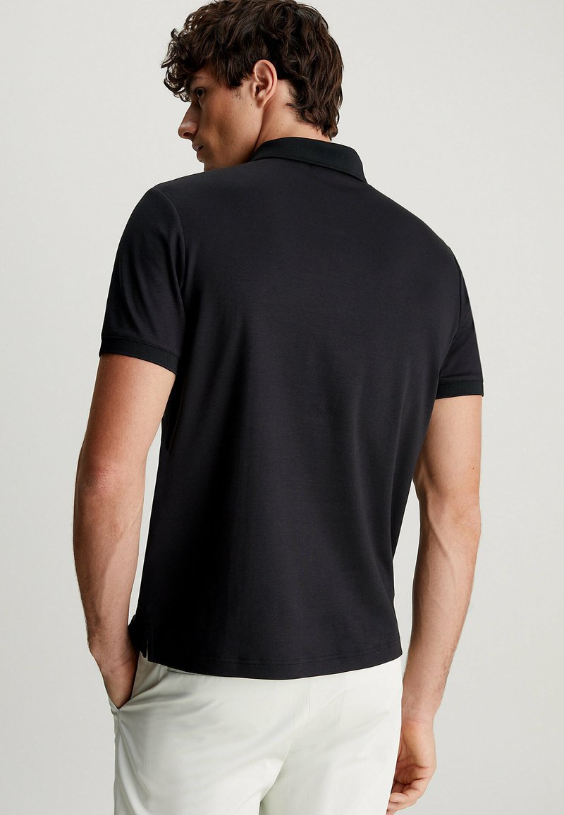 Zwarte polo met korte mouwen, een kraag en een rechte zoom, gecombineerd met lichte broek. Gladde textuur en minimaal ontwerp.
