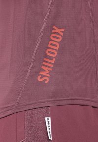 Top atletico color marrone caratterizzato da un tessuto strutturato, cuciture a punto e il nome del marchio "SMILODOX" in rosso bold sul lato.