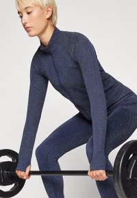Navy Sport-Outfit mit einem taillierten, strukturierten Design, ausgestattet mit einem Reißverschluss und langen Ärmeln, während eine Hantel in einer Hebepose gehalten wird.