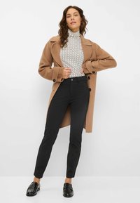 Brauner Wollmantel, gemusterter Rollkragenpullover in Schwarz und Weiß, enger schwarzer Hose und glänzenden schwarzen Slippern. Glatte Texturen und taillierte Passform.