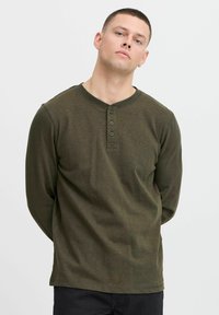 Olivenfarbendes Langarm-Henley-Shirt mit Rundhalsausschnitt, Knopfleiste mit drei Knöpfen und feiner Textur. Das Model steht mit verschränkten Händen.