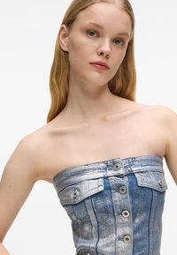 Νεαρή γυναίκα με ανοιχτόχρωμο δέρμα και ξανθά μαλλιά, φορώντας ένα μεταλλικό μπλε τοπ από τζιν χωρίς straps, με κουμπιά και τσέπες.