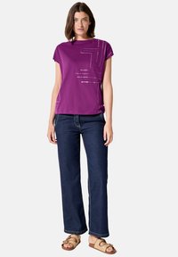 Camiseta morada de manga corta con un patrón geométrico plateado, combinada con pantalones vaqueros de pierna ancha en denim oscuro y sandalias marrones.
