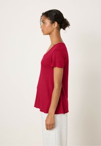 Raudonas trumpomis rankovėmis viršus su V-įpjovimu ir laisvu siluetu, pagamintas iš lengvos medžiagos, minkštai krentantis ties kraštu, kuriant atsitiktinį įvaizdį.